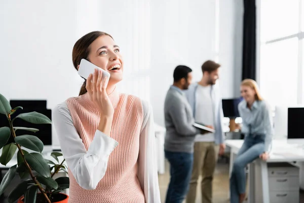 Mujer de negocios sonriente hablando en el teléfono inteligente cerca de colegas en el fondo borroso en la oficina - foto de stock
