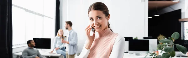 Smiling businesswoman looking at camera while talking on smartphone in office, banner — Stock Photo