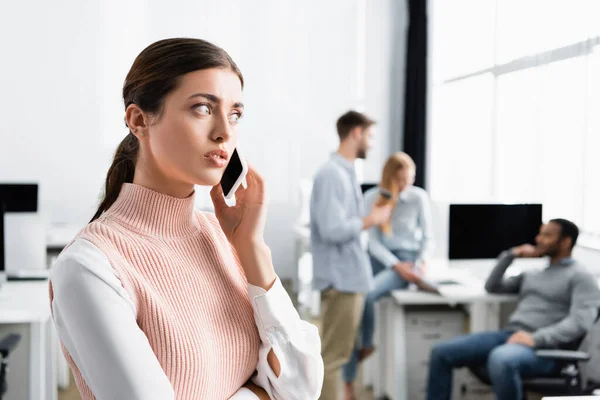 Young businesswoman talking on smartphone with colleagues on blurred background in office — Stock Photo