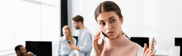 Mujer de negocios que tiene idea mientras habla en el teléfono inteligente cerca de colegas en fondo borroso, pancarta - foto de stock