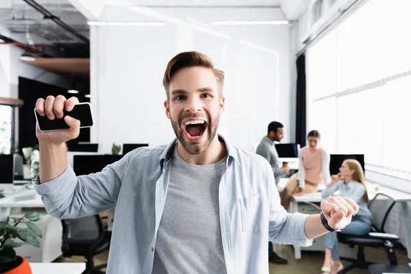 Fröhlicher Geschäftsmann hält Smartphone in der Hand, während er in die Kamera neben Kollegen auf verschwommenem Hintergrund blickt — Stockfoto