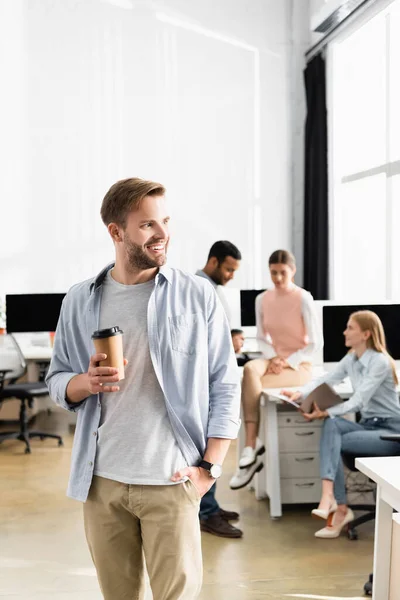 Улыбающийся бизнесмен держит кофе, чтобы подойти к многонациональным коллегам на размытом фоне в офисе — стоковое фото