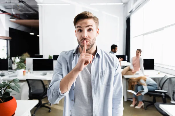 Young businessman showing shh gesture near colleagues on blurred background in office — Stock Photo