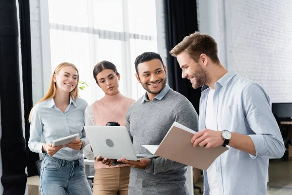 Hombre de negocios sonriente sosteniendo una carpeta de papel cerca de colegas multiculturales con computadora portátil y tableta digital - foto de stock