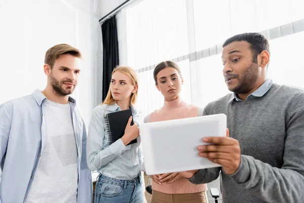 Indian businessman holding digital tablet near colleagues with notebook in office — Stock Photo