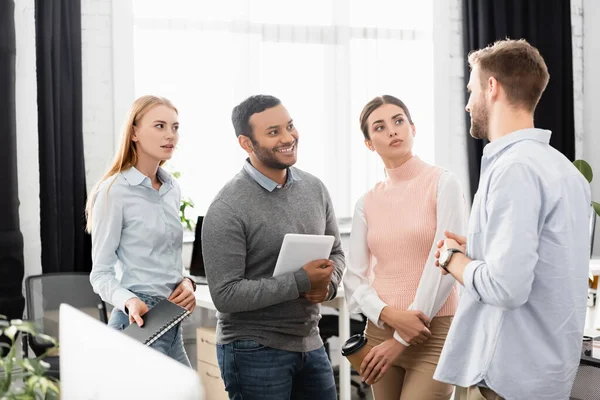 Empresario hablando con sonrientes empresarios multiétnicos sosteniendo tableta digital y café para ir en la oficina - foto de stock