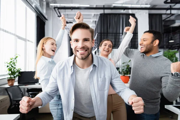 Smiling businessman showing yeah gesture near cheerful multiethnic colleagues on blurred background in office — Stock Photo