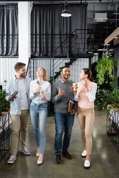 Cheerful multicultural businesspeople with paper folder and coffee to go talking while walking in office — Stock Photo