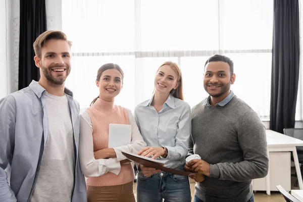 Multiethnic businesspeople with paper folder and digital tablet smiling at camera in office — Stock Photo