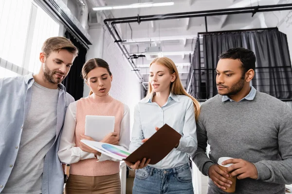 Businesswoman holding paper folder with colorful swatches near multiethnic colleagues with digital tablet and coffee to go in office — Stock Photo
