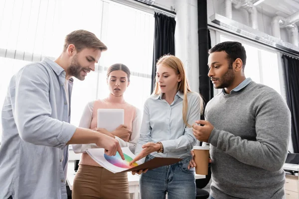 Jeunes hommes d'affaires multiethniques avec café à emporter et tablette numérique pointant vers des échantillons colorés au bureau — Photo de stock