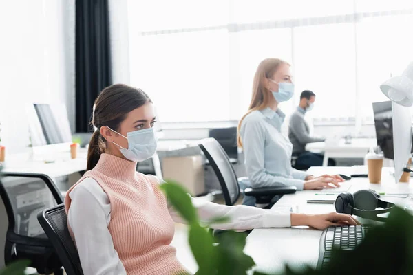 Geschäftsfrau in medizinischer Maske sitzt neben Computertastatur und Kollegen auf verschwommenem Hintergrund — Stockfoto