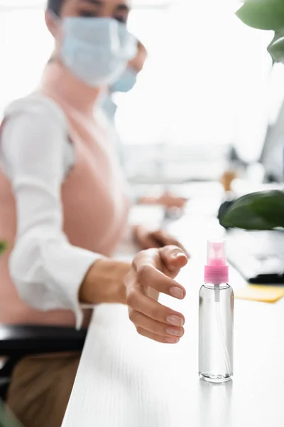 Бизнесмен в медицинской маске тянет руку к дезинфицирующему средству на столе на размытом фоне — стоковое фото