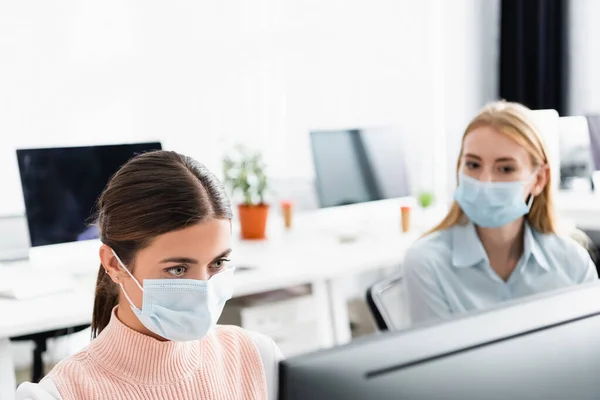 Empresaria en máscara médica mirando monitor de computadora cerca de colega en la oficina - foto de stock