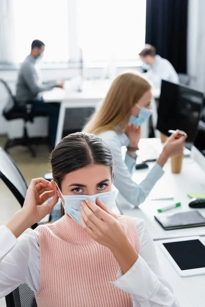 Geschäftsfrau blickt in Kamera, während sie auf verschwommenem Hintergrund medizinische Maske neben Kollegin trägt — Stockfoto