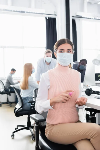 Femme d'affaires dans le masque médical à l'aide d'un désinfectant pour les mains tout en collègues travaillant sur fond flou — Photo de stock