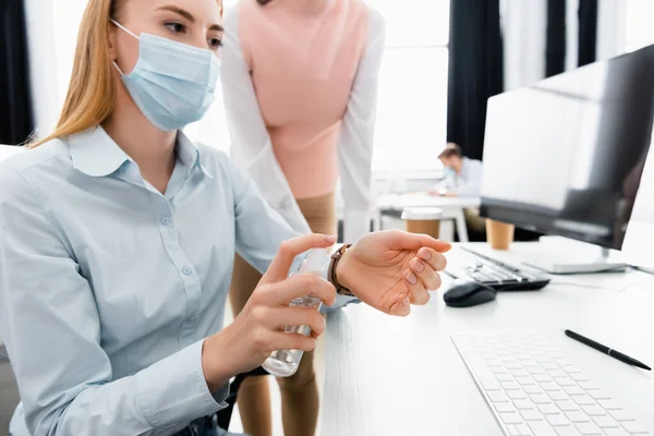 Donna d'affari in maschera medica utilizzando disinfettante mano vicino collega e computer su sfondo sfocato — Foto stock