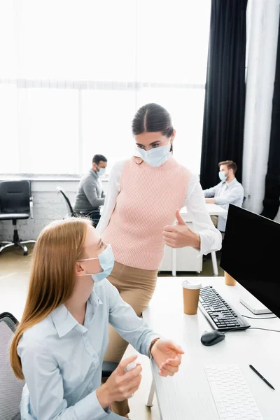Empresaria en máscara médica mostrando como colega cercano usando desinfectante de manos en la oficina - foto de stock