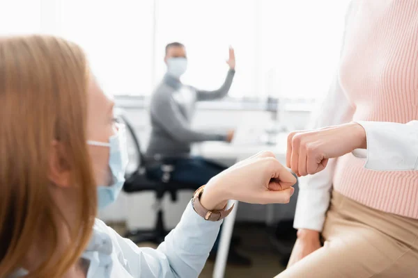 Femme d'affaires dans le masque médical faisant coup de poing avec un collègue sur fond flou — Photo de stock