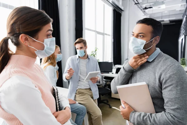 Indian businessman in medical mask touching neck while talking with colleague on blurred foreground in office — Stock Photo