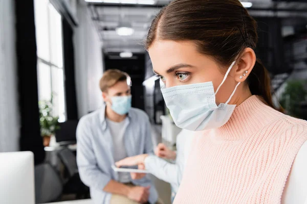 Femme d'affaires en masque médical debout près de collègues sur fond flou dans le bureau — Photo de stock