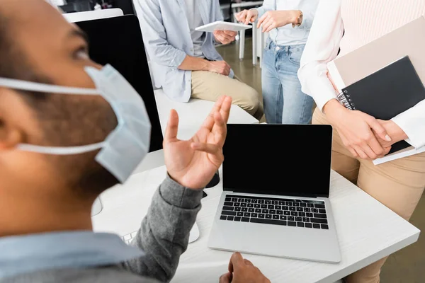 Empresarios con tableta digital y portátil de pie cerca de colega indio en máscara médica en la oficina - foto de stock