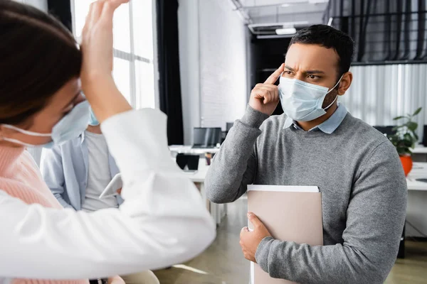 Indian businessman with paper folder pointing at head near sick colleague on blurred foreground in office — Stock Photo