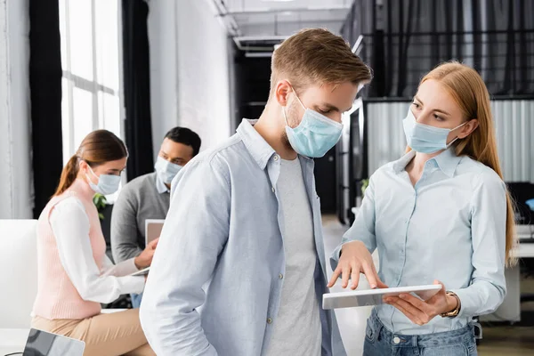 Businesswoman in medical mask using digital tablet near colleague in office — Stock Photo