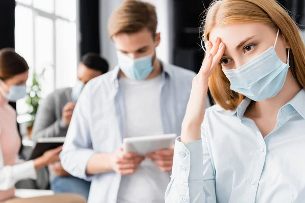 Хвора бізнес-леді в медичній масці торкається голови біля колег на розмитому фоні в офісі — стокове фото