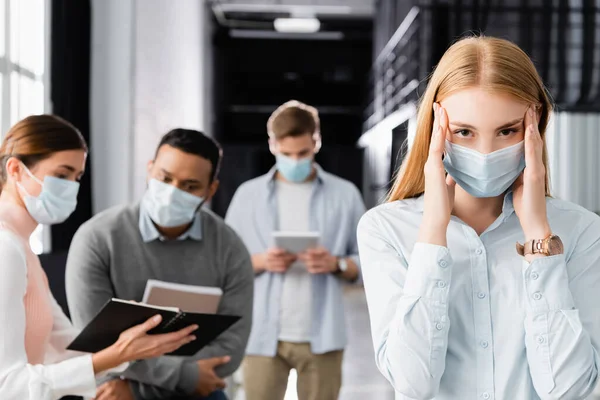 Хворі бізнес-леді в медичній масці торкаються близько, в той час як багатоетнічні колеги працюють на розмитому фоні — стокове фото
