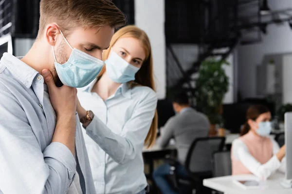 Kranker Geschäftsmann mit medizinischer Maske berührt vor verschwommenem Hintergrund Kollegin am Hals — Stockfoto