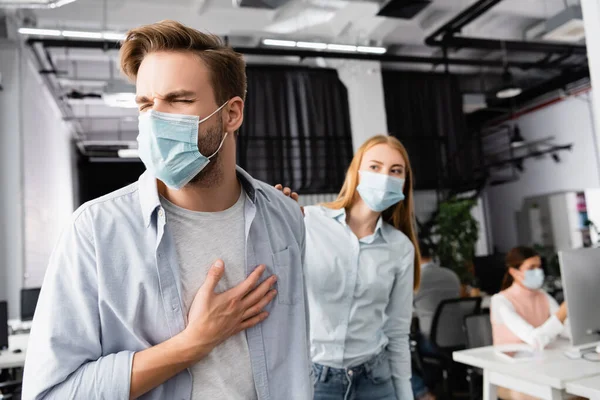 Empresario enfermo en máscara médica tocando el pecho cerca de colega en fondo borroso en la oficina - foto de stock