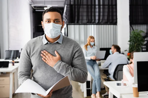 Indischer Geschäftsmann mit medizinischer Maske berührt Brust, während er Papiermappe im Büro hält — Stockfoto
