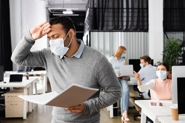 Enfermo empresario indio en máscara médica sosteniendo carpeta de papel cerca de colegas en fondo borroso en la oficina - foto de stock