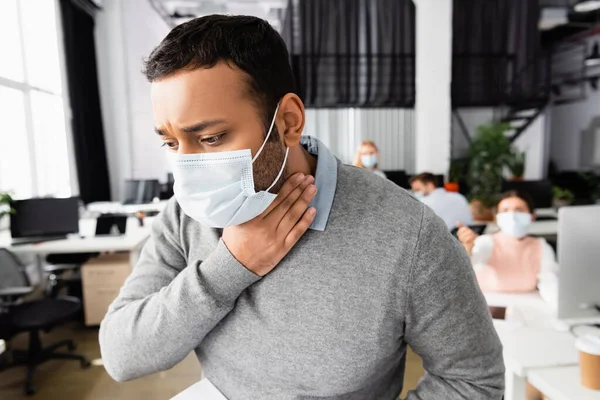 Kranker indischer Geschäftsmann mit medizinischer Maske berührt Hals im Büro — Stockfoto