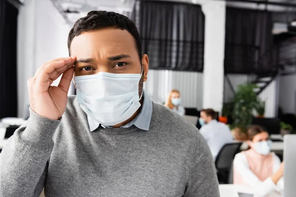 Ill indian businessman in medical mask looking at camera near colleagues on blurred background — Stock Photo