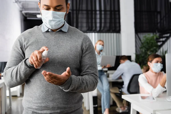 Uomo d'affari indiano in maschera medica utilizzando disinfettante mano in bottiglia mentre i colleghi che lavorano su sfondo sfocato — Foto stock