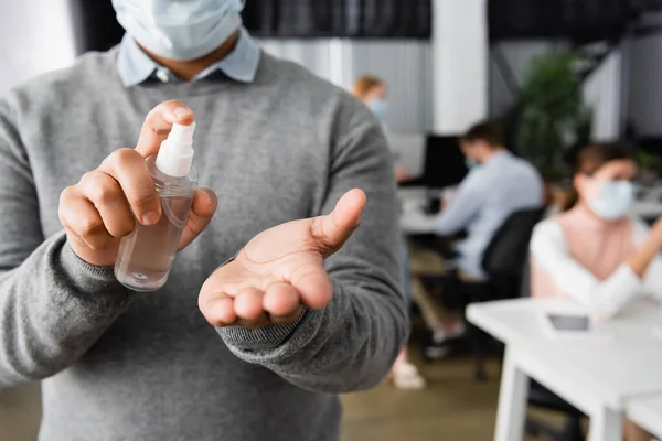 Vista recortada de hombre de negocios en máscara médica sosteniendo botella de desinfectante de manos en la oficina - foto de stock