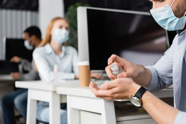 Ausgeschnittene Ansicht des Geschäftsmannes in medizinischer Maske mit Händedesinfektionsmittel in der Nähe von Computern und Kollegen auf verschwommenem Hintergrund — Stockfoto