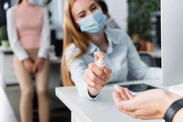 Empresaria en mascarilla médica rociando desinfectante de manos a mano de colega en primer plano borroso en la oficina - foto de stock