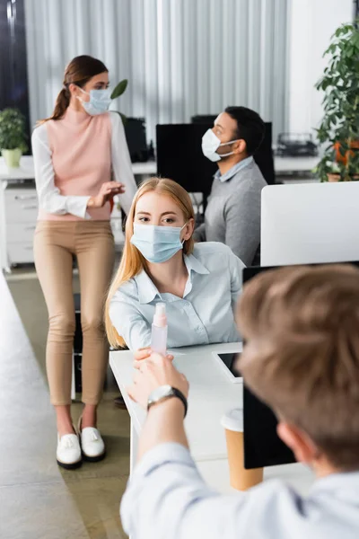 Businesswoman in medical mask giving hand sanitizer to colleague on blurred foreground while working in office — Stock Photo