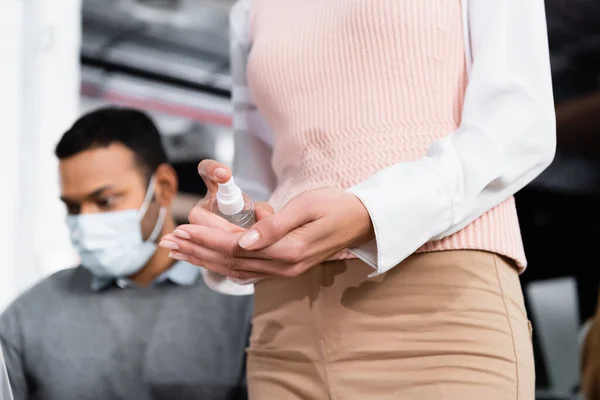 Vista recortada de la mujer de negocios usando desinfectante de manos cerca de colega indio en máscara médica sobre fondo borroso - foto de stock