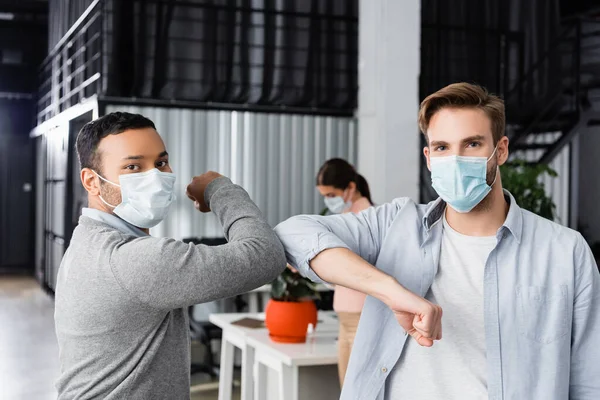 Multicultural businessmen in medical masks giving high five with elbows in office — Stock Photo