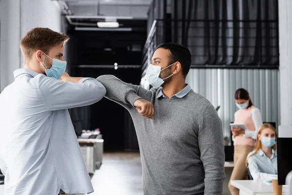 Empresários multiétnicos em máscara médica dando alta cinco com cotovelos perto de colegas em fundo turvo no escritório — Fotografia de Stock