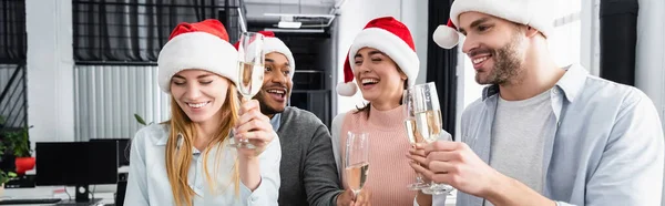 Imprenditori multiculturali sorridenti in cappelli di Babbo Natale che tengono bicchieri di champagne in ufficio, striscione — Foto stock