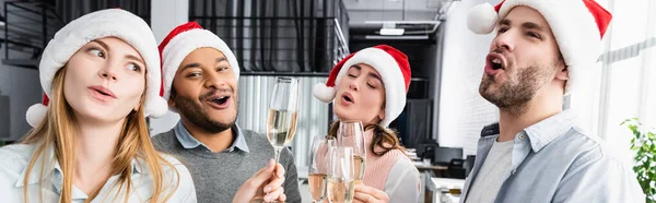 Des hommes d'affaires multiethniques excités dans des chapeaux de Père Noël tenant des verres de champagne au bureau, bannière — Photo de stock