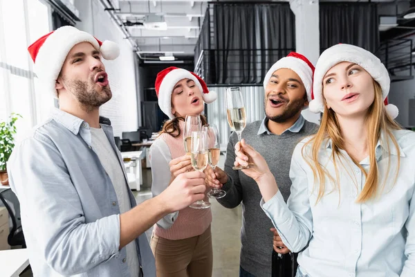 Imprenditori multiculturali eccitati in cappelli di Babbo Natale che tengono bicchieri di champagne in ufficio — Foto stock