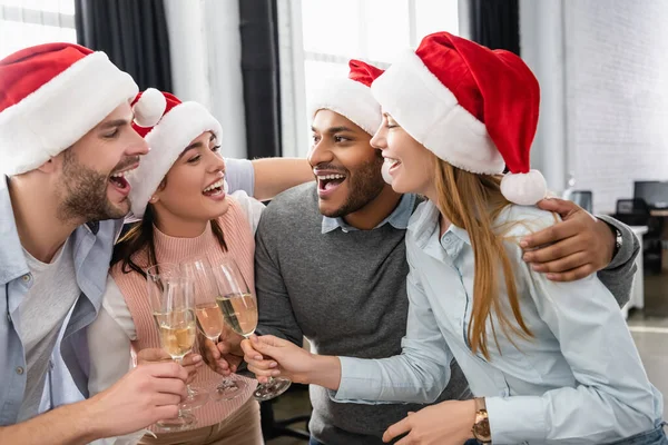 Empresários multiétnicos em chapéus de Papai Noel abraçando enquanto clica com champanhe no escritório — Fotografia de Stock