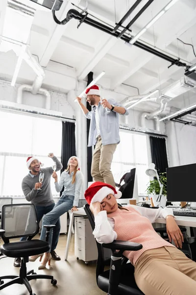 Empresaria durmiendo en sombrero de santa cerca de alegres colegas multiétnicos con champán sobre fondo borroso - foto de stock