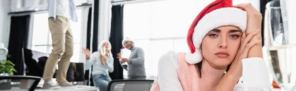 Donna d'affari stanca in cappello di Babbo Natale guardando la macchina fotografica vicino a vetro di champagne e colleghi su sfondo sfocato, banner — Foto stock
